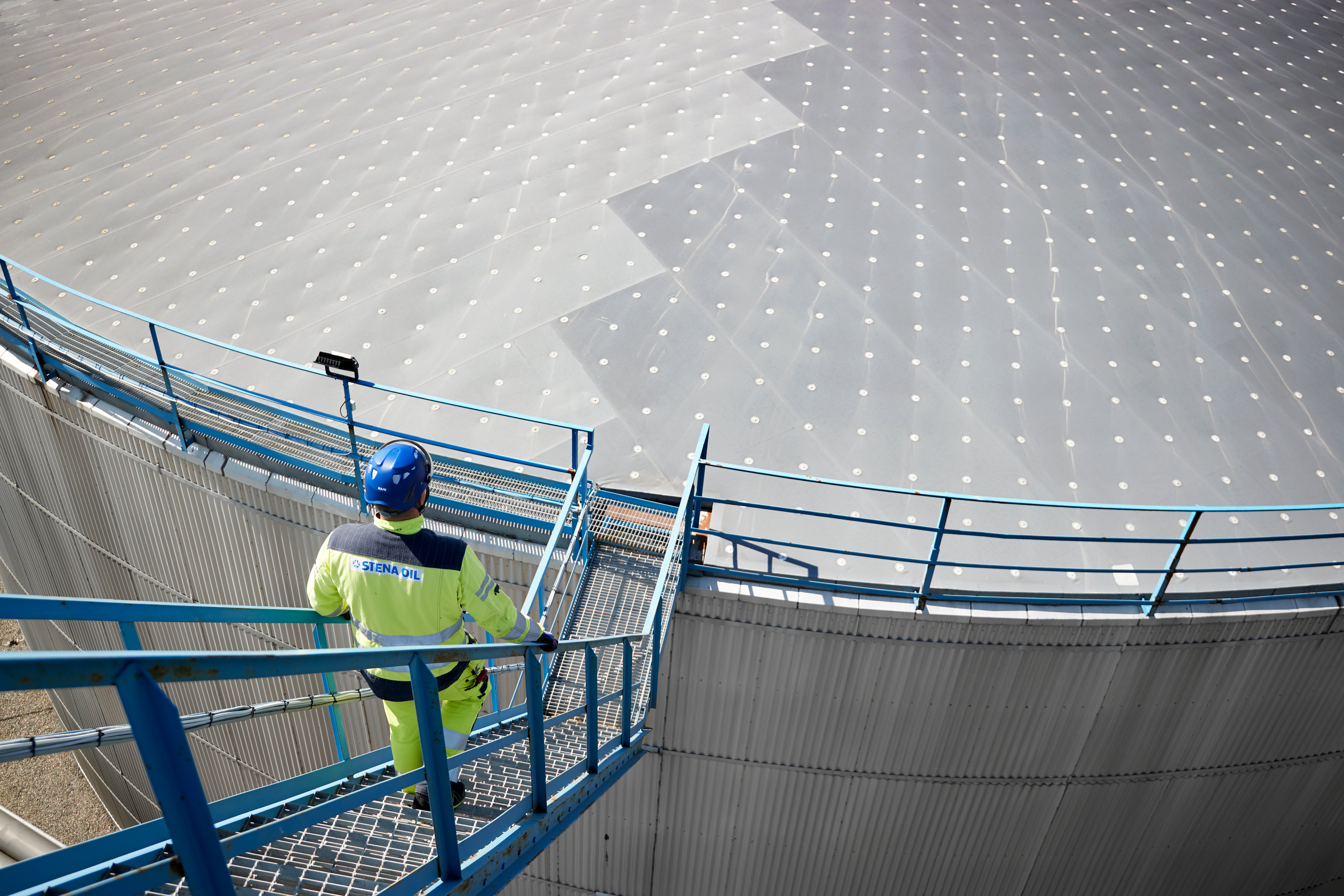 Man in yellow jacket and construction helmet walking down stairs to an oil plant